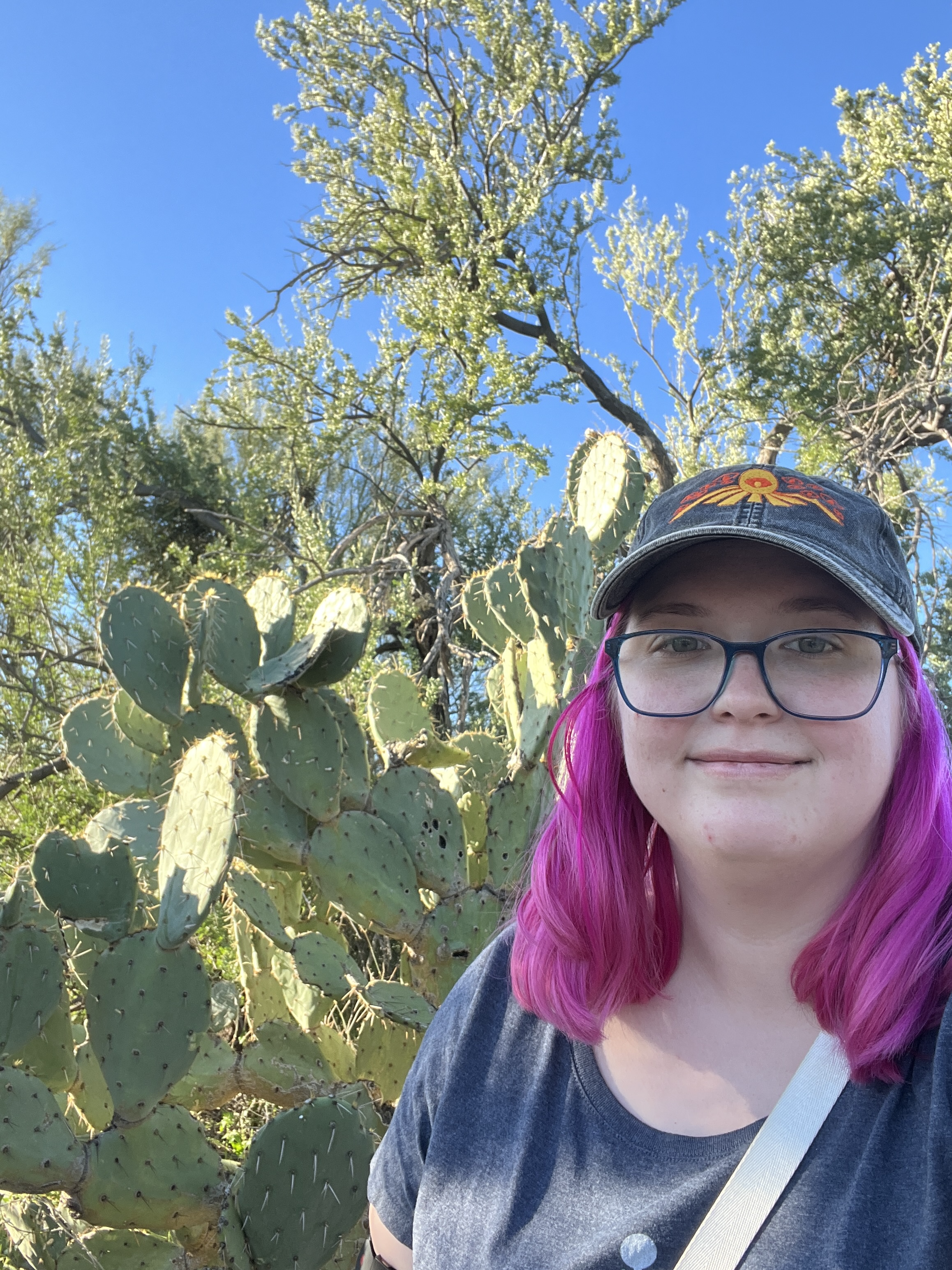 Sarah, a white person with shoulder length pink hair, wearing a baseball cap and standing in front of a prickly pear cactus
